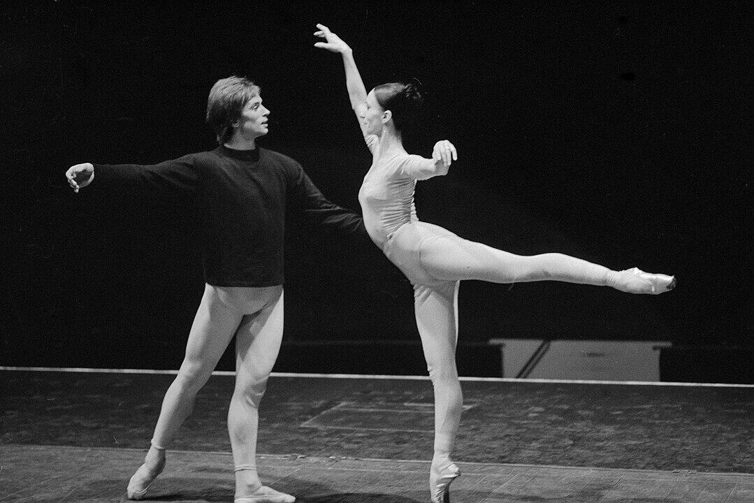 A male ballet dancer in a black sweater supports a female dancer in a graceful pose, showcasing their practice in a dim studio.