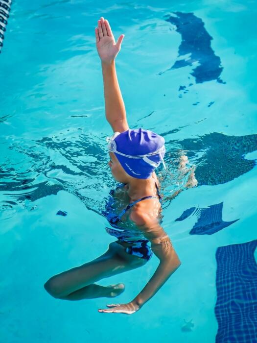 A young swimmer with a blue cap raises one hand above water, surrounded by bright blue pool tiles and clear water.