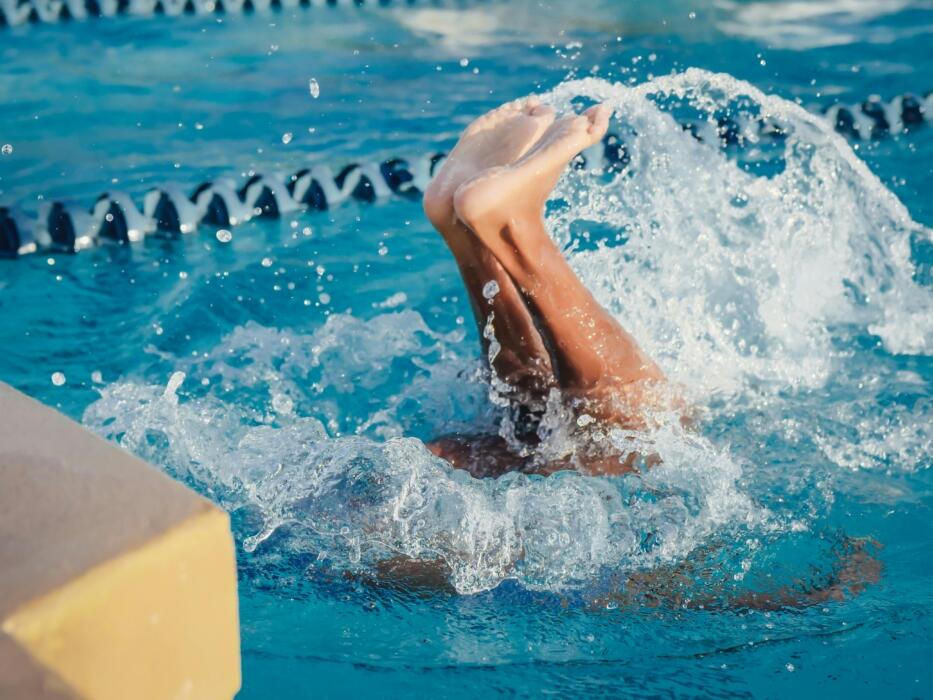 A person with their legs raised high, creating splashes in a vibrant blue swimming pool water.