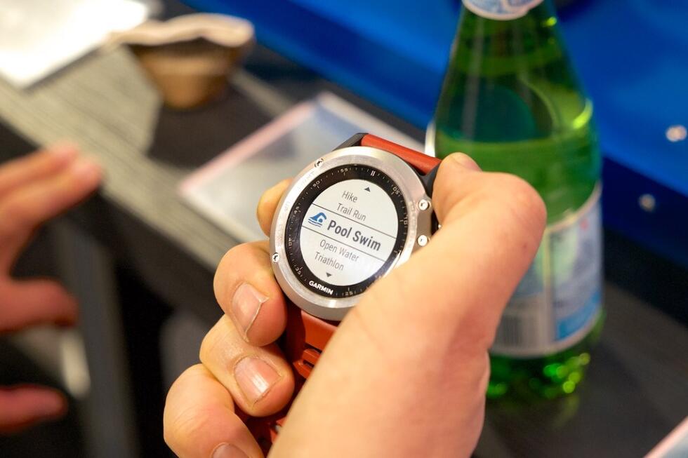 A hand holding a Garmin watch displaying the "Pool Swim" option, with a bottle of sparkling water in the background.