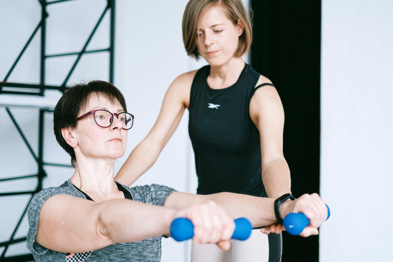 A person performs an exercise with dumbbells, guided by a trainer in a gym setting. Both individuals are focused and engaged.