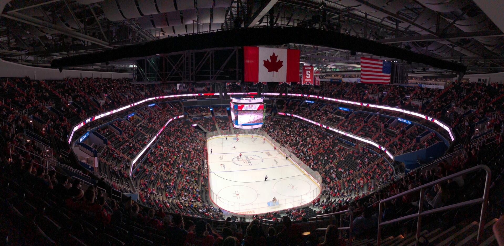 A panoramic view of a packed ice hockey arena, featuring flags of Canada and the USA, with players on the ice and fans in red attire.