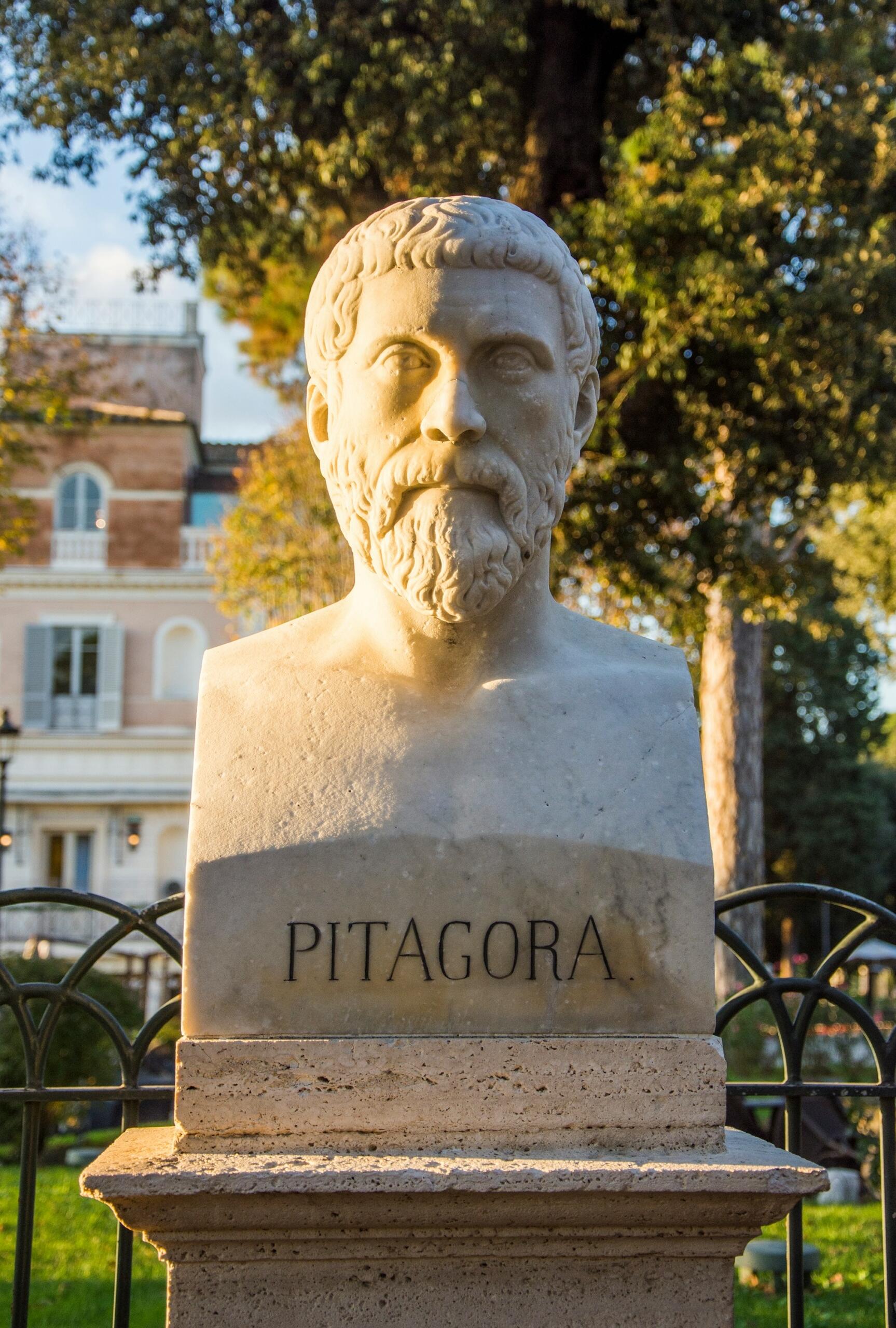Marble bust of Pythagoras on a pedestal in a sunlit garden with trees and a building in the background.