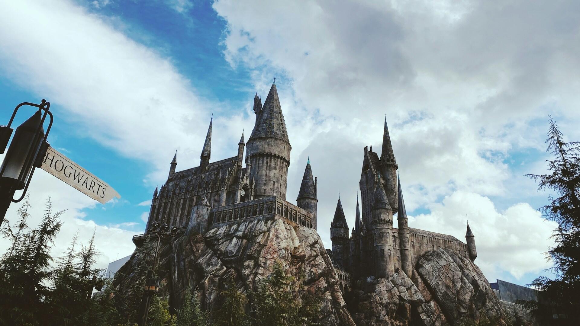 View of Hogwarts castle on a rocky hill under a partly cloudy sky with a "Hogwarts" sign on the left and evergreen trees in the foreground.