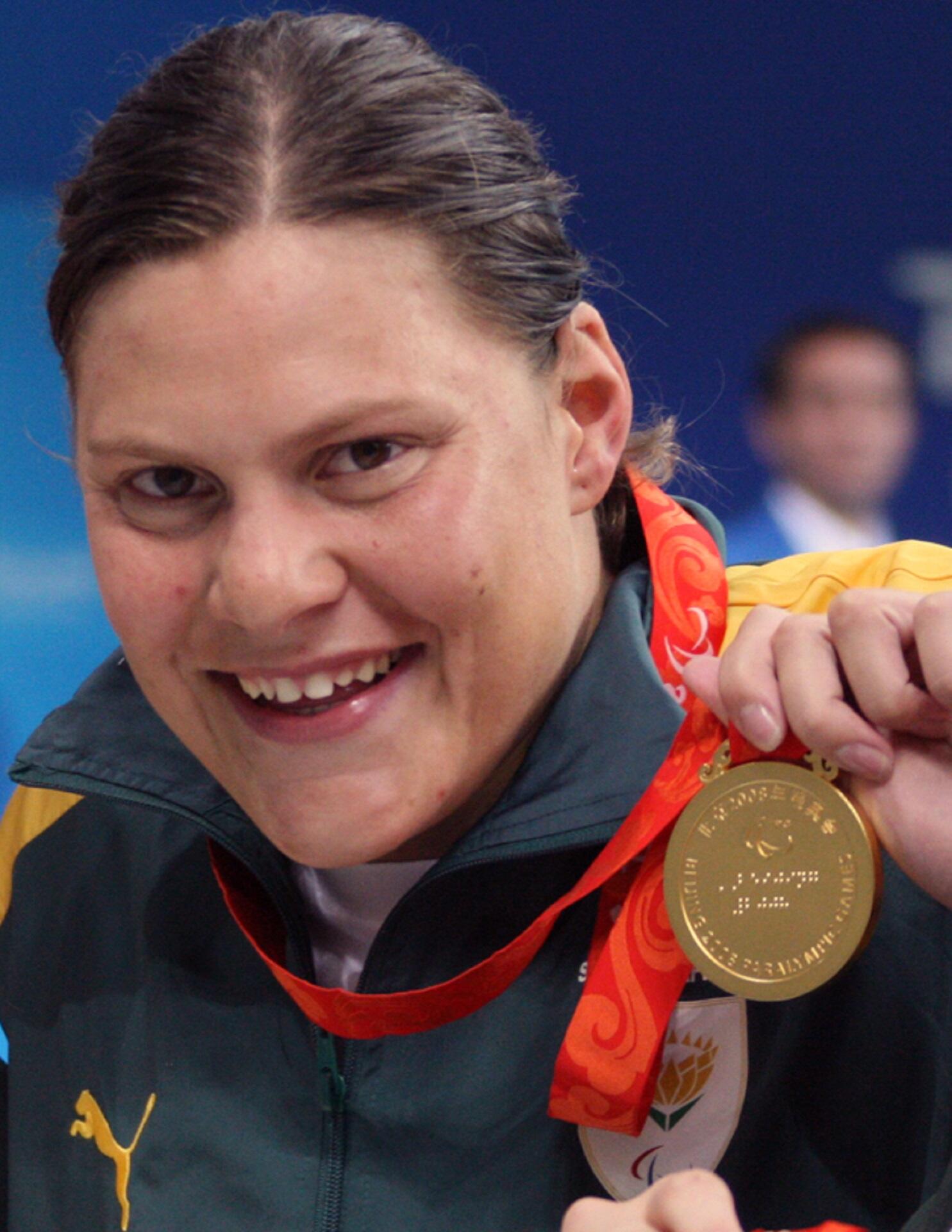 A woman with her hair pulled back smiles as she holds a gold medal, mounted on an orange lanyard, close to her face.