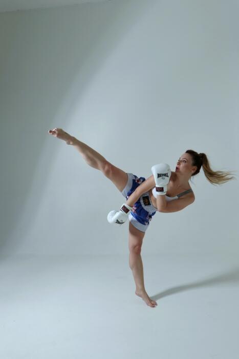 A woman wearing athletic clothing and boxing gloves practices kickboxing in a bright, all-white training room. Source: Skoro Photo.